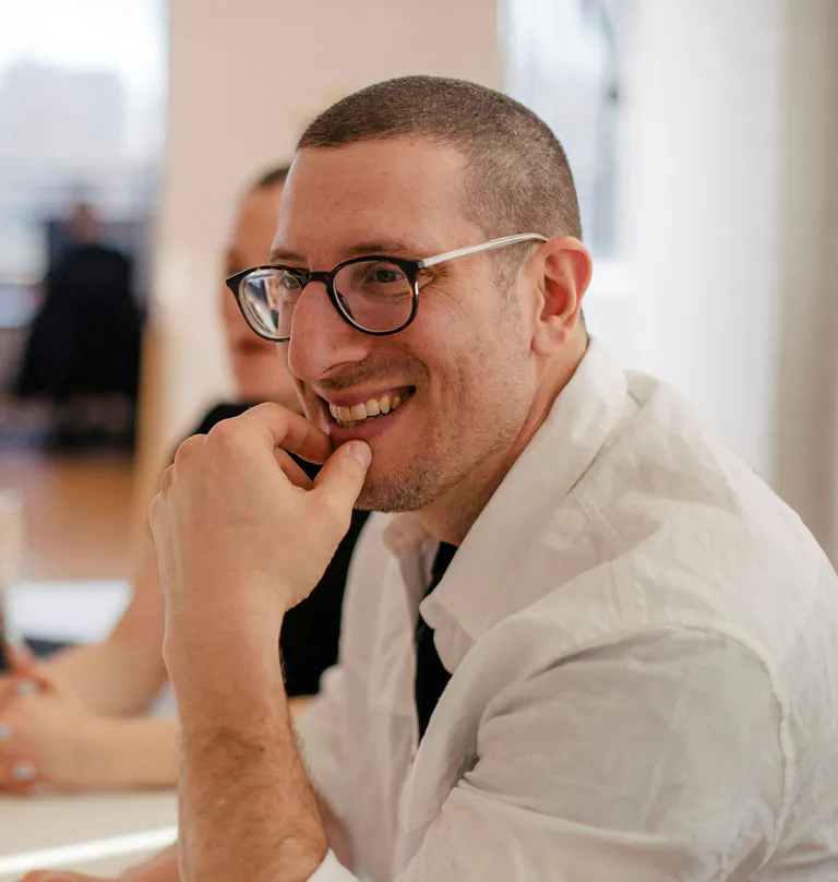 A designer in glasses smiling at the studio table, a colleague and a laptop beside him