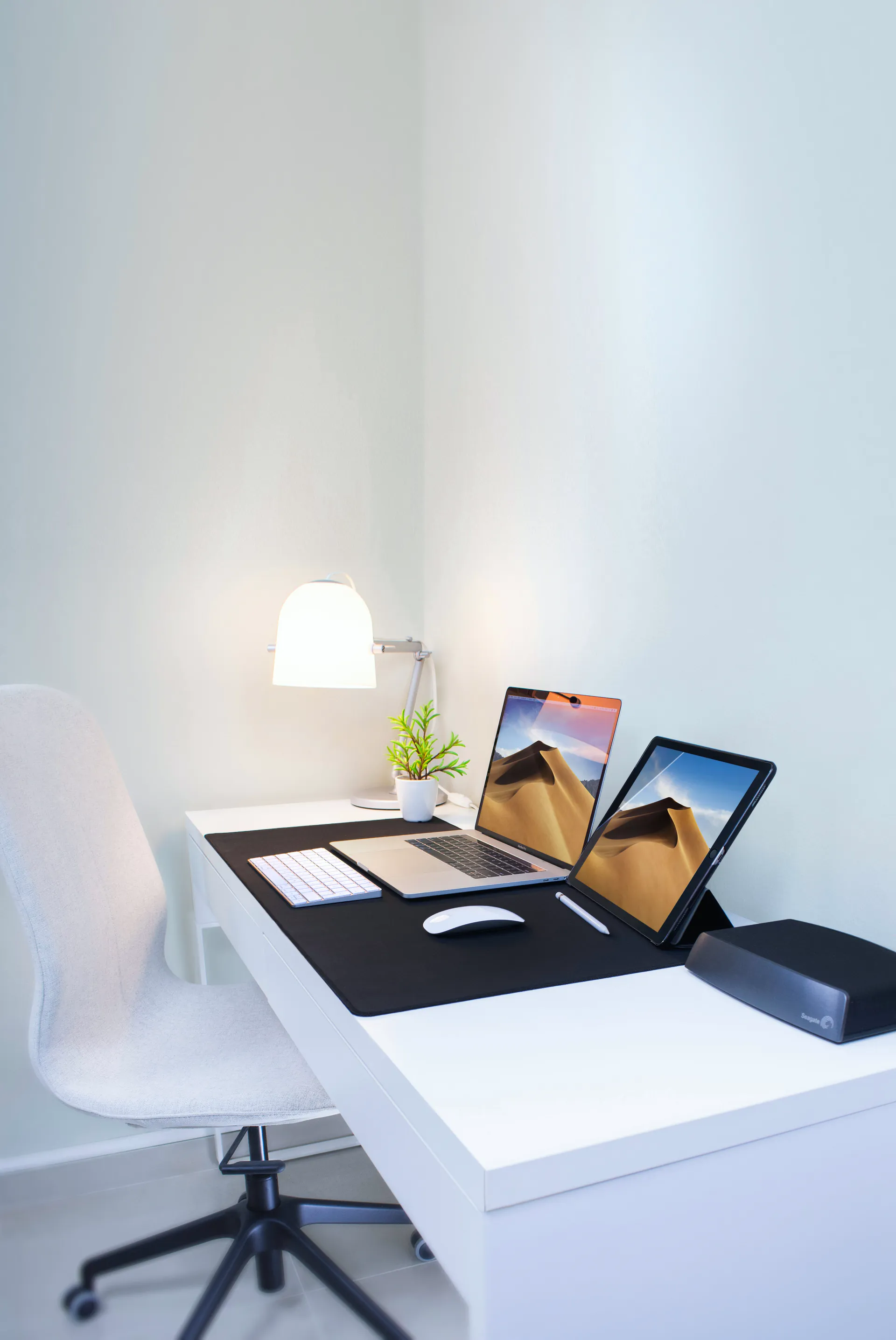 A white desk with a laptop and a tablet set up in the bright corner of an office