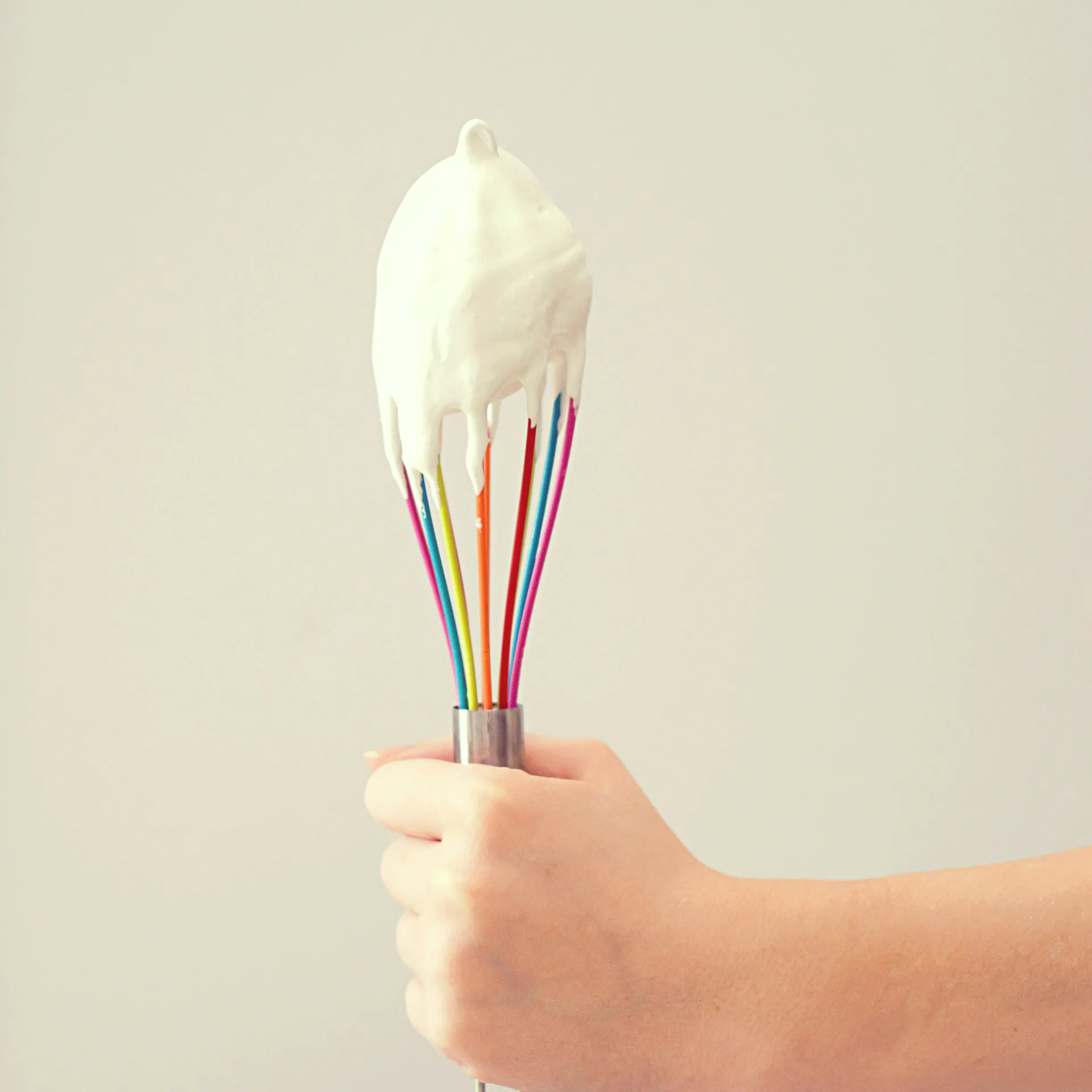 A hand holding up a rainbow-coloured whisk dipped in meringue