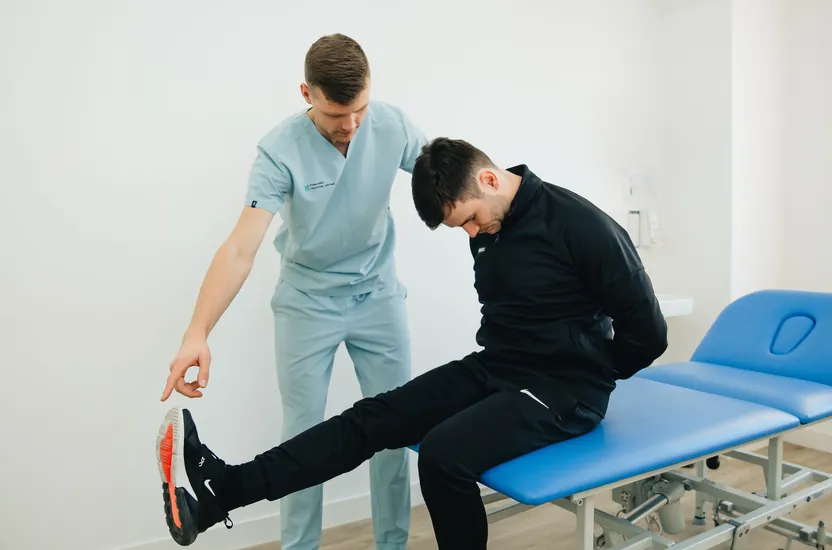 A physiotherapist in blue scrubs guiding a patient through a hamstring stretch on a treatment table