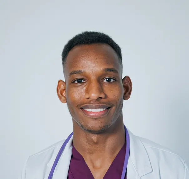 A smiling young doctor in a white coat over maroon scrubs, a stethoscope round his neck
