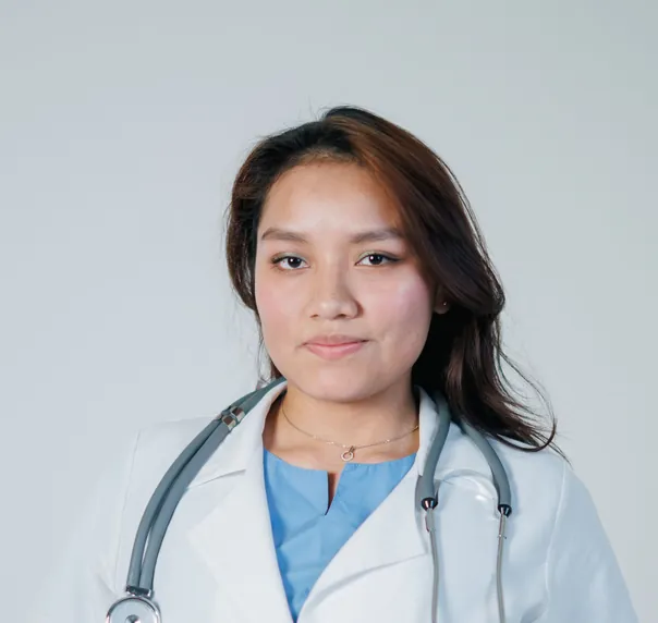 A doctor with long dark hair in a white coat over light blue scrubs, a stethoscope round her neck