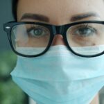 A close-up of a doctor in black-rimmed glasses and a blue surgical mask, looking into the camera