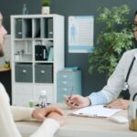 A doctor in glasses and a white coat taking notes as a bearded patient talks to her across the desk
