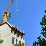 A yellow tower crane working over the top floor of a new concrete apartment block, framed by trees against a blue sky