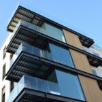 Balconies with glass balustrades and dark steel soffits stacked up a pale apartment building with a strip of timber cladding, against a blue sky