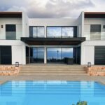 A white two-storey house with glass balconies above a stone terrace whose wide steps lead down to a swimming pool