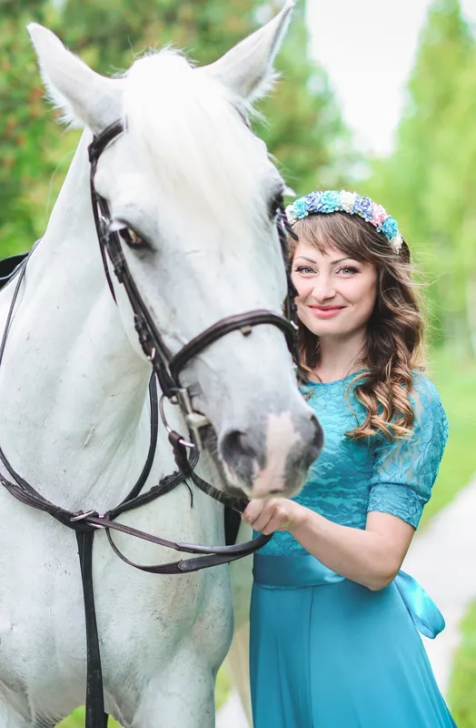 A woman in a blue dress with a garland of flowers, holding the bridle of a grey horse