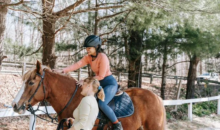 An instructor steadying a chestnut horse as a rider in a helmet settles into the saddle, among pine trees