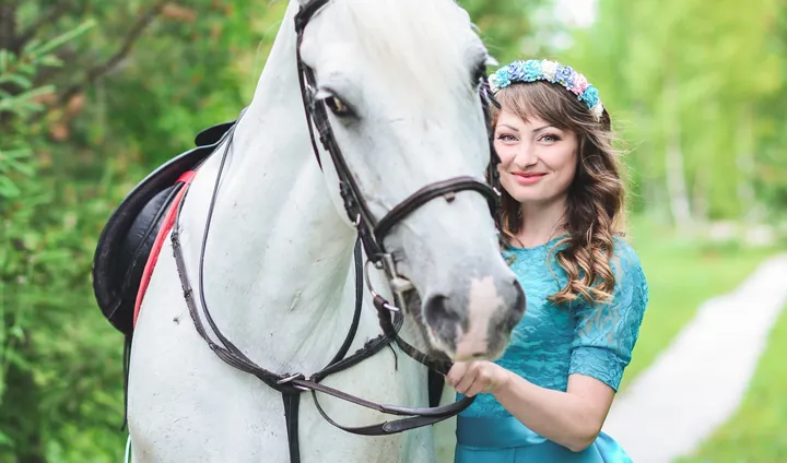 A woman in a blue dress holding the bridle of a grey horse on a woodland path