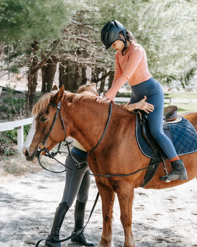 A rider in a helmet swinging into the saddle of a chestnut horse while an instructor holds it