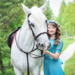 A smiling woman in a blue dress and a flower crown holding the bridle of a grey horse on a woodland path
