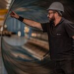 An engineer in a hard hat standing beside the open end of a large steel pipe section in a fabrication hall