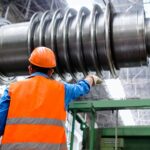 An engineer in an orange hard hat and hi-vis vest pointing along a huge machined shaft in a workshop