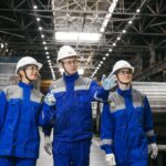 Three engineers in blue overalls and white hard hats walking and talking along a steel mill aisle