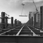 Rows of steel columns and beams of a new building frame, a load swinging from a crane, in black and white