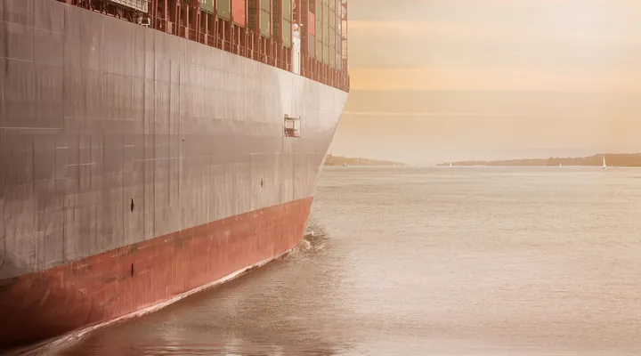 The bow of a loaded container ship moving through calm water at sunset