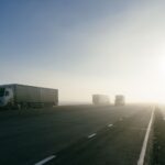 Two articulated trucks with curtain-sided trailers on an empty highway in morning fog, two more further back