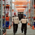 Two warehouse workers in beige and brown overalls carrying cardboard boxes along an aisle of orange pallet racking