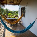 A wooden balcony with two hammocks, one blue and one yellow, a table and chairs, and green trees beyond the railing