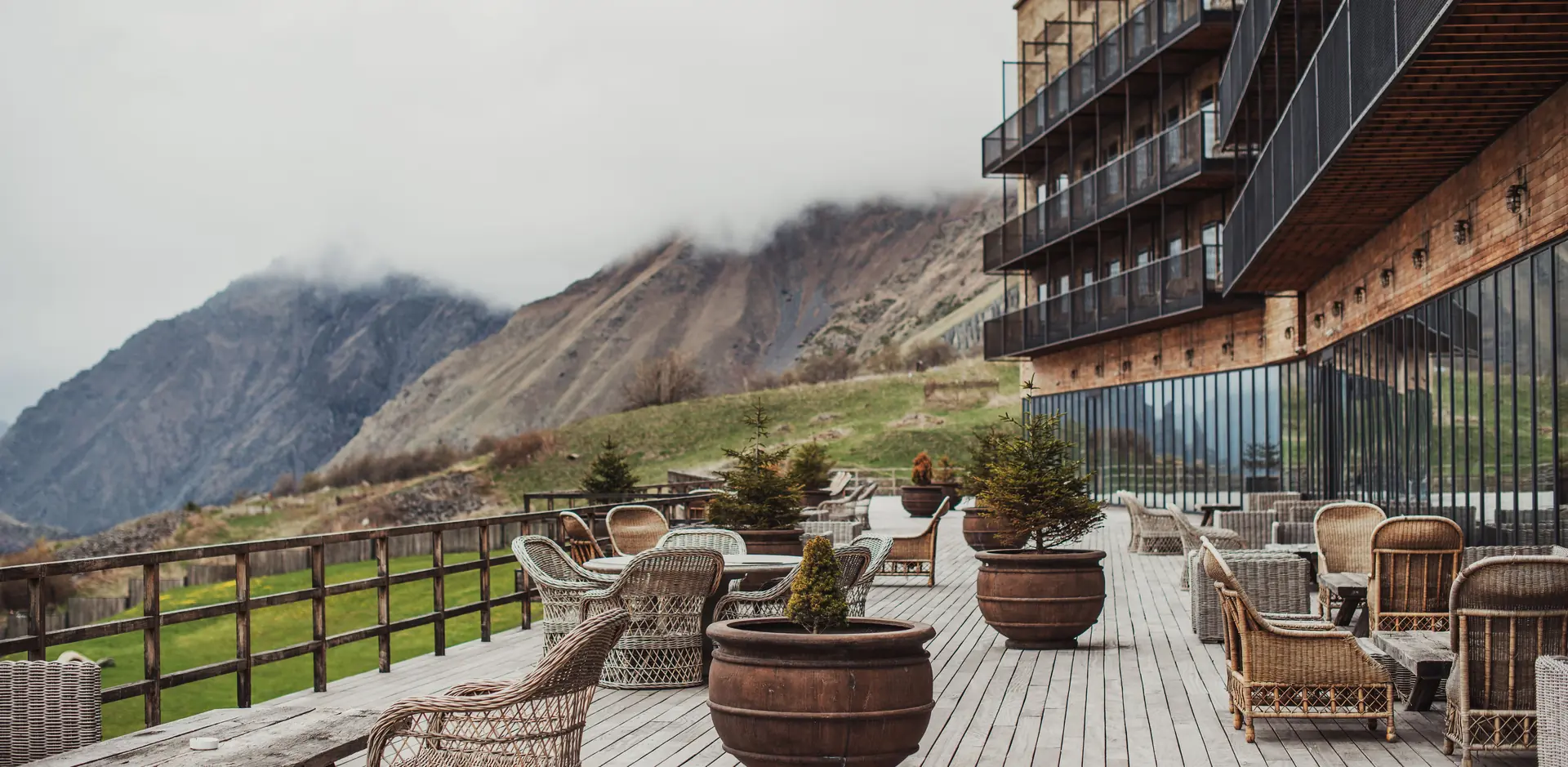 Wicker armchairs and potted pines on a long wooden terrace beside a hotel, clouds on the mountains