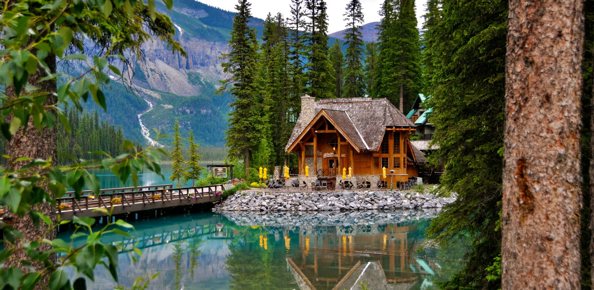 A timber lodge on a stony point of a turquoise lake, framed by tall pines and a mountain