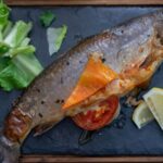 A whole roasted trout on a slate board with lettuce, a slice of tomato and lemon wedges