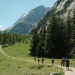 A line of walkers on a grassy path up a green valley, pines and a rocky ridge above them