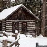A snow-covered log cabin in a wood, a snowy split-rail fence in front of it