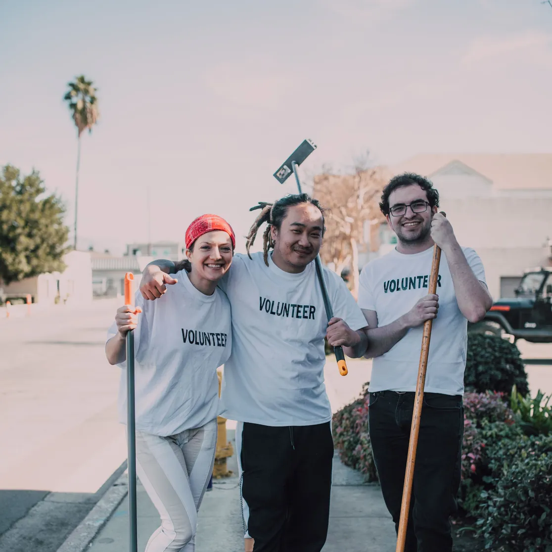 Three volunteers in white T-shirts, arms around each other, holding a broom and a mop on a sunny sidewalk