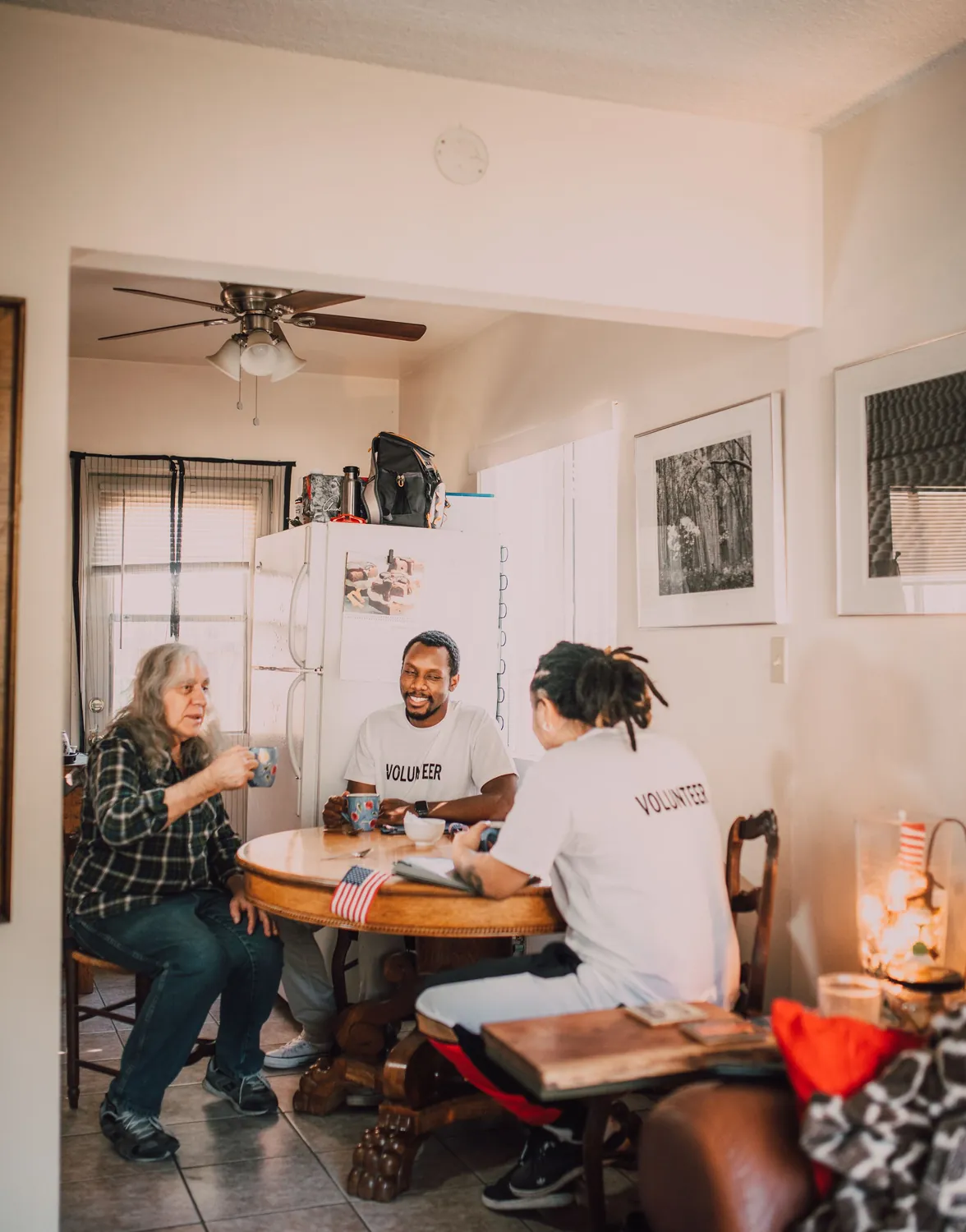An older woman in a plaid shirt at a round table with two volunteers, in a bright front room with a ceiling fan
