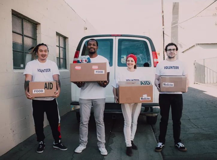 Four volunteers in white VOLUNTEER T-shirts outside a van, holding boxes labelled Food, Aid and Medicine