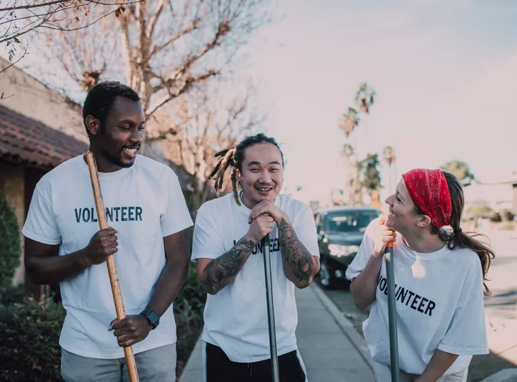 Three volunteers with brooms laughing together on a sunny sidewalk