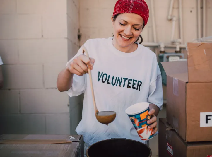 A volunteer in a red bandana ladling soup into a paper cup beside boxes marked Food