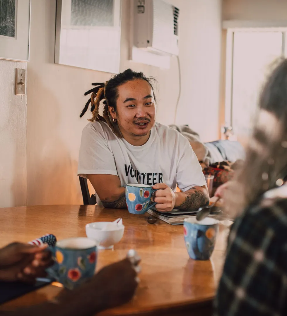 A volunteer with dreadlocks smiling across a kitchen table set with painted coffee mugs
