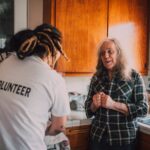 A volunteer in a white VOLUNTEER T-shirt talking with an older woman in her kitchen