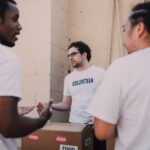Three volunteers in white VOLUNTEER T-shirts talking over a box marked Food outside the pantry