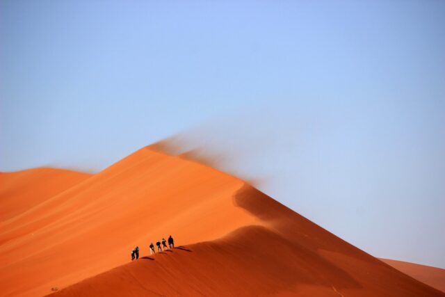 A red sand dune with a line of tiny figures walking along the crest
