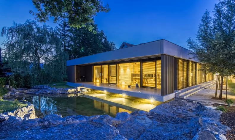 A low glass house lit up at dusk beside a rock pool, with trees behind
