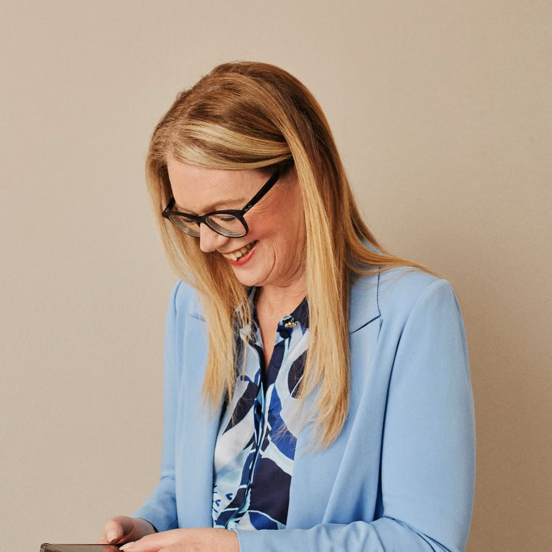 Maria Williams, smiling in glasses and a pale blue jacket as she reads her phone