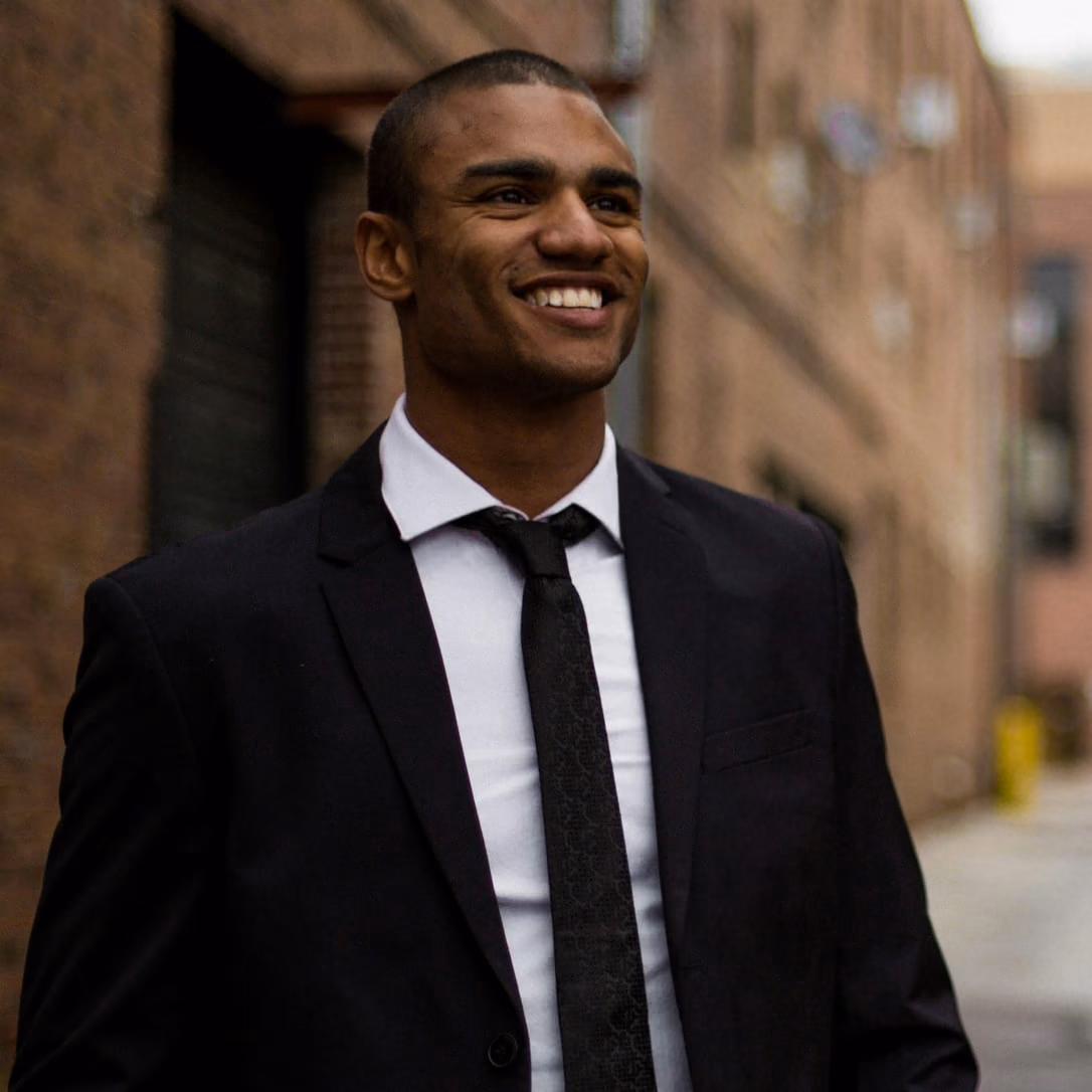Patrick Joe, smiling in a black suit and tie on a city street