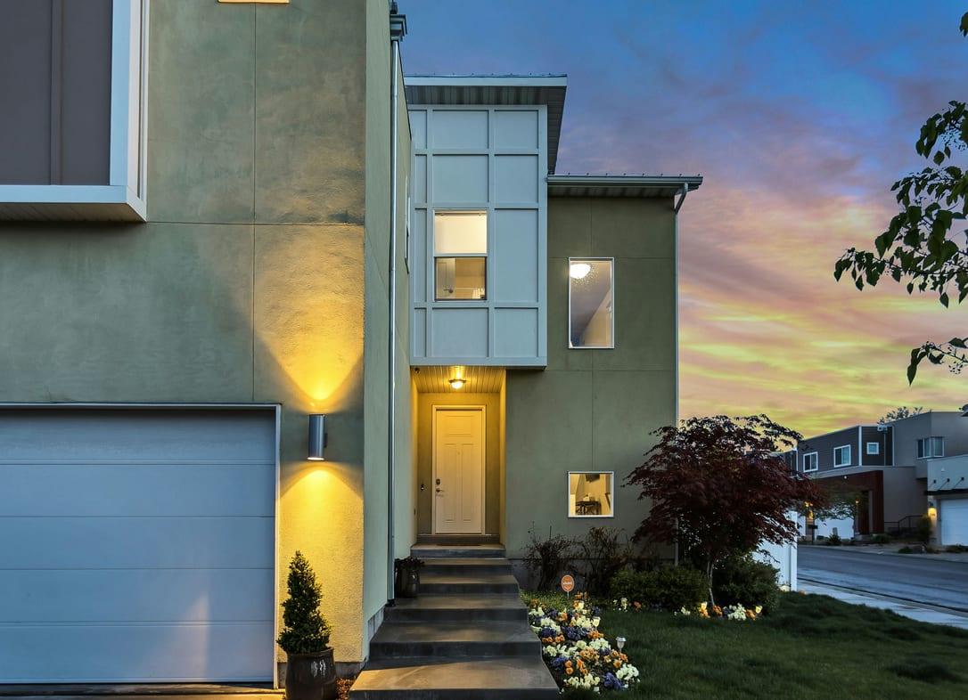 Contemporary townhouse with grey render, a garage door and warm wall lights at dusk