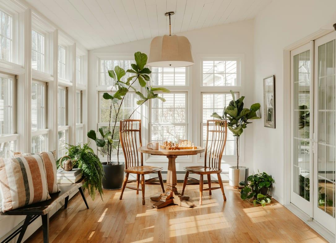 Sunny dining corner with house plants, a round table, wooden chairs and tall white windows