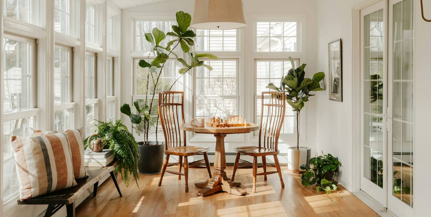 Sunny dining corner with house plants, a round table, wooden chairs and tall white windows
