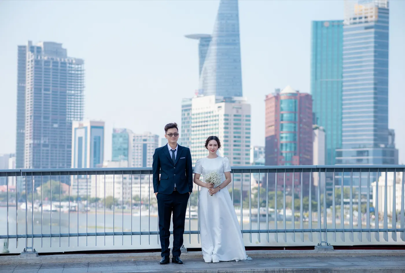 Mai and Khoa standing at the railing of the bridge with the river and the city skyline behind them