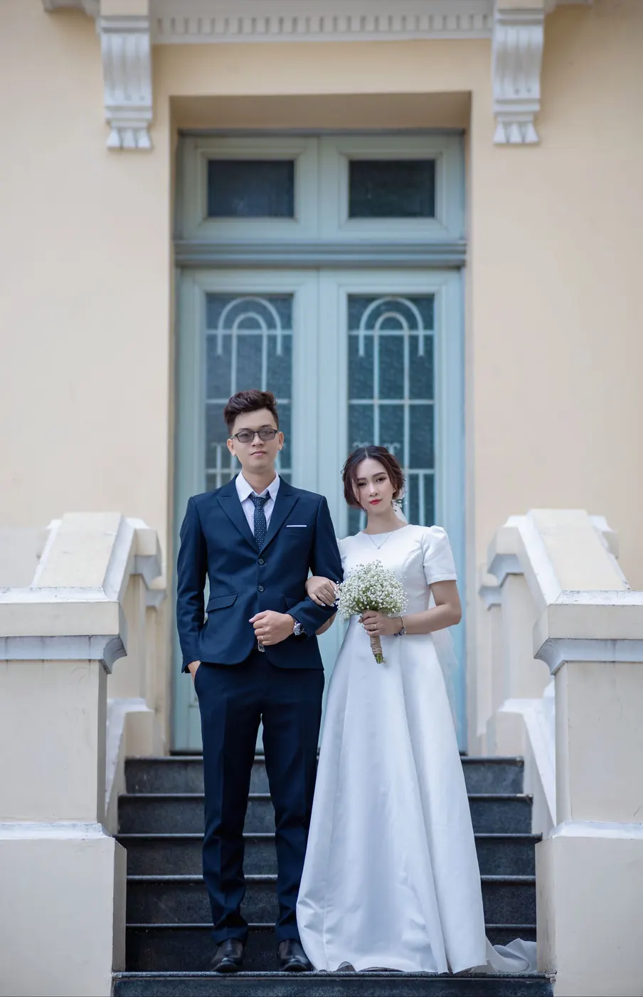 The couple on the steps of an old cream-painted building, arm in arm