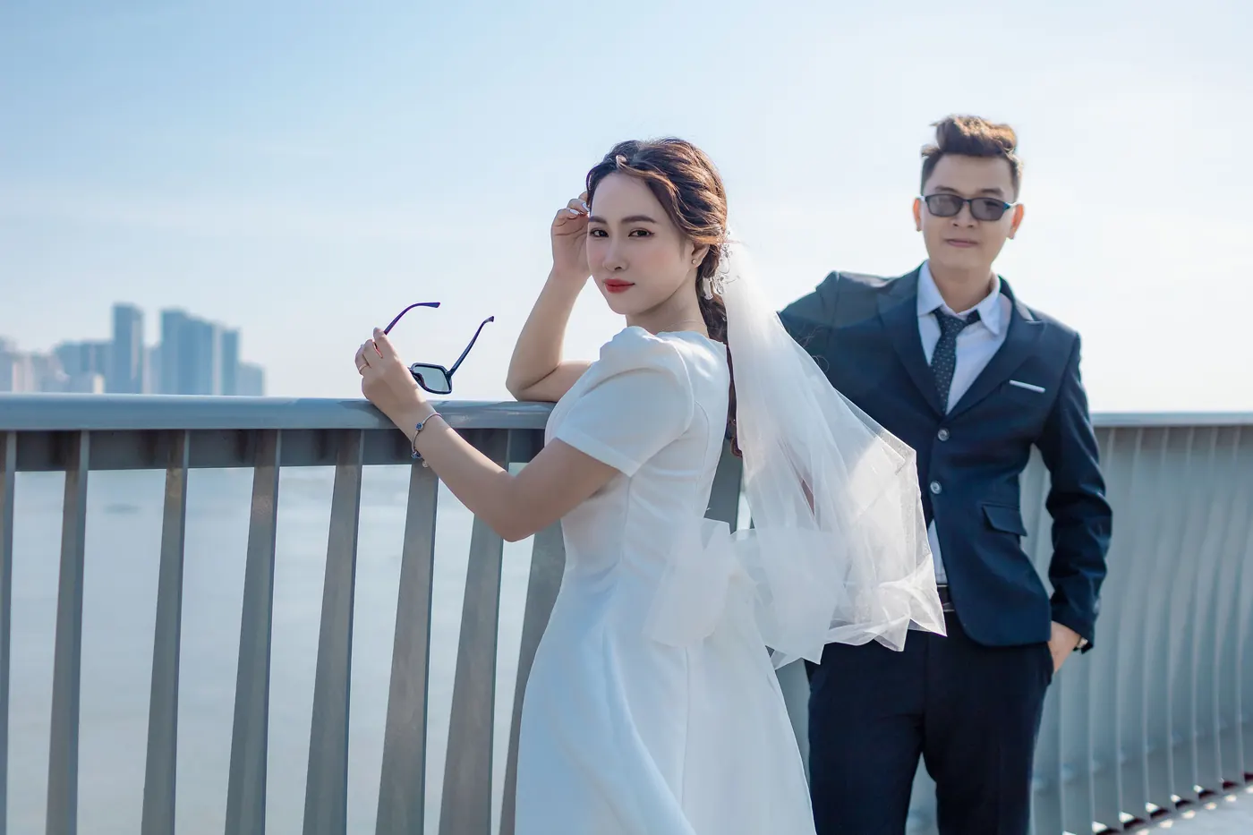 Mai holding her sunglasses and leaning on the railing, Khoa beside her with a hand in his pocket