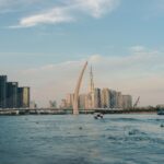 The white arch of a cable-stayed bridge over a wide river at dusk, a speedboat below and tall apartment towers behind
