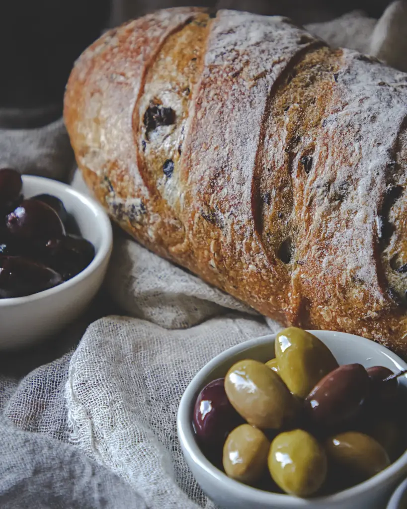 A dark seeded loaf, sliced, with olives beside it