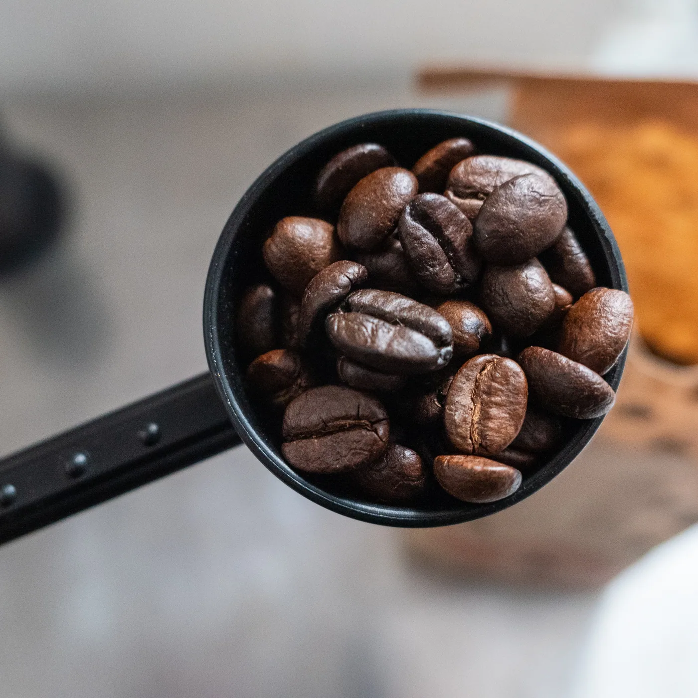 A black scoop brimming with roasted coffee beans on a warm tan surface